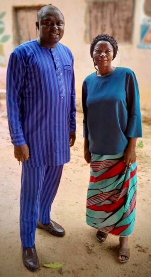 Minna, widow of Rev. Samuel Ahmed, stands alongside a male companion outside a building. She's dressed in a blue top and a patterned skirt of red, green, and white, complemented by brown sandals. The man on her left is clad in a blue-striped traditional outfit with black shoes. The backdrop features a weathered wall and door. Minna continues her devotion to serving the Lord by teaching children and women, and actively goes door-to-door in villages to share the Gospel.