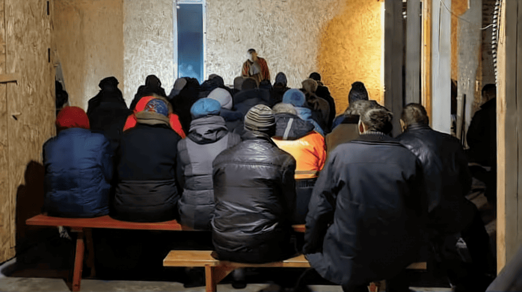 A group of people, dressed in winter attire, seated on wooden benches in an indoor setting with rustic, bare plywood walls, attentively listening to a person standing in front of them.