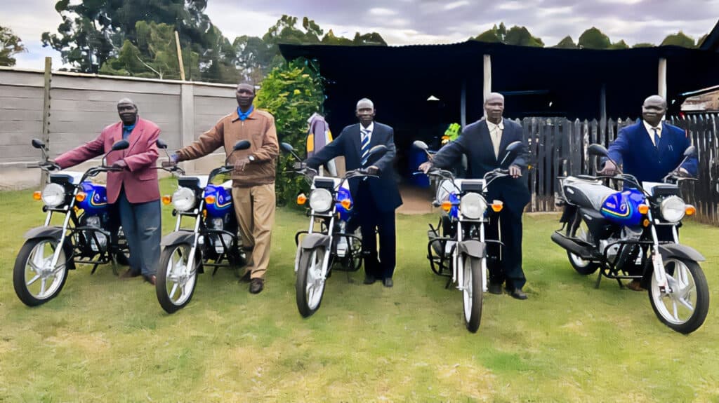 A group of five men standing proudly beside motorcycles in an open field, with trees and a wooden fence in the background. Each man is dressed in formal attire and positioned next to a motorcycle.