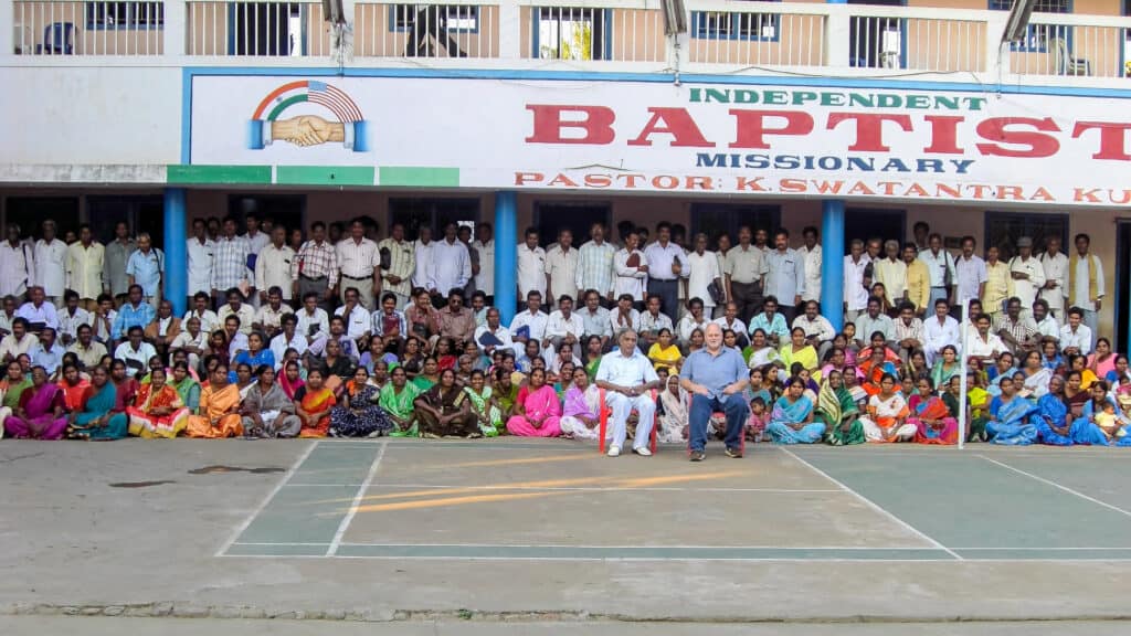 A large group of individuals of Indian ethnicity, dressed predominantly in traditional Indian attire and vibrant sarees, assembled in rows, and two individuals, one Indian and one White, seated in the forefront. The group is in front of a 2-story building labeled "INDEPENDENT BAPTIST MISSIONARY". Next to the building's label there is a logo featuring hands clasped under a rainbow with the colors of the Indian flag.