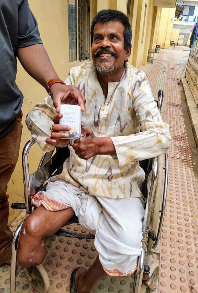 A blind man in a wheelchair with a cheerful expression, wearing a patterned shirt and white trousers, being handed an audio Bible device by another individual outside a yellow building, with a corridor in the background.