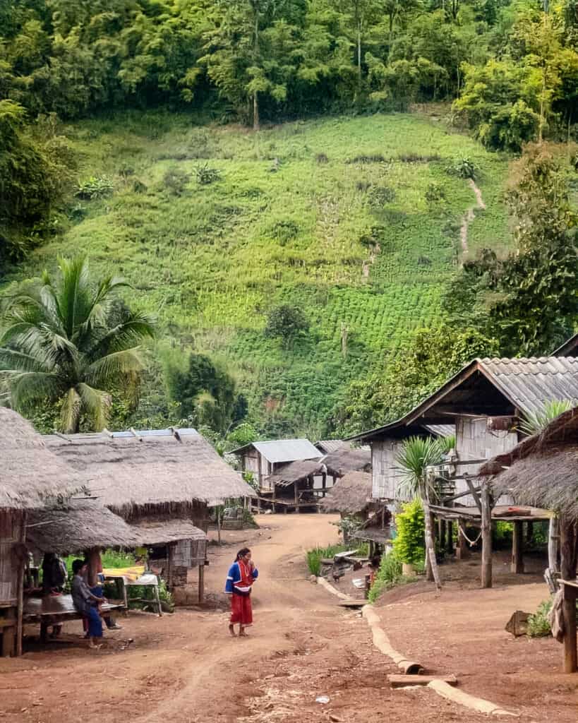 A tranquil village nestled among lush green hills, with traditional thatched-roof huts. In the foreground, a woman in vibrant blue and red clothing stands in the middle of a dirt path, with other villagers visible around the huts.