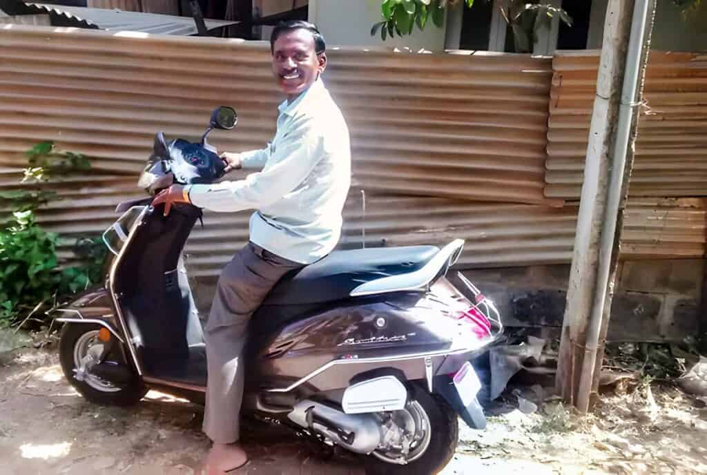 A man with a broad smile is sitting on a black scooter parked outdoors. He is wearing a light-colored long-sleeved shirt and dark trousers. The scooter has a glossy finish with pink and white accents and the word "Activa" written on the side. The background shows a corrugated metal sheet fence and some foliage, suggesting a sunny day in a residential or semi-urban area.