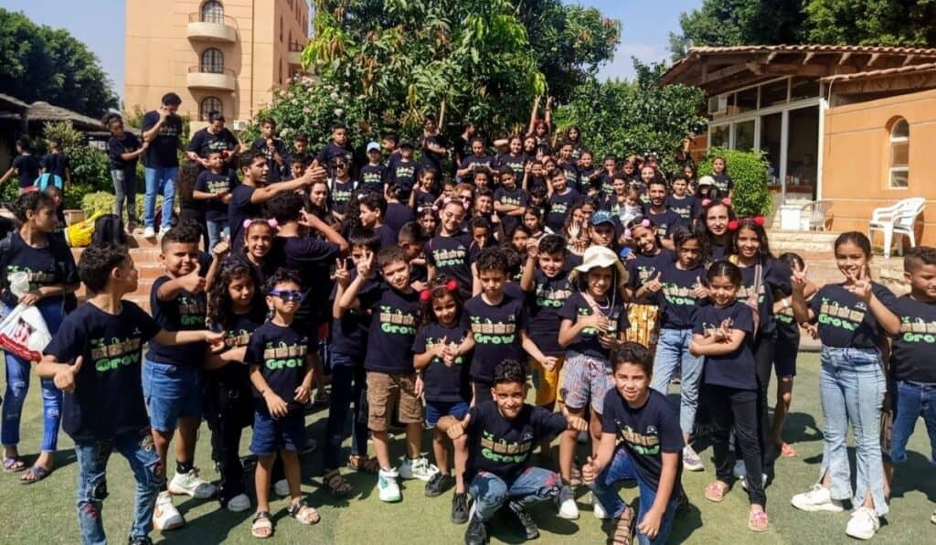 A large group of children and a few adults wearing matching black t-shirts with the word "Grow" printed on them, posing outdoors in front of a building and garden. Many of the children are making playful gestures and expressions.
