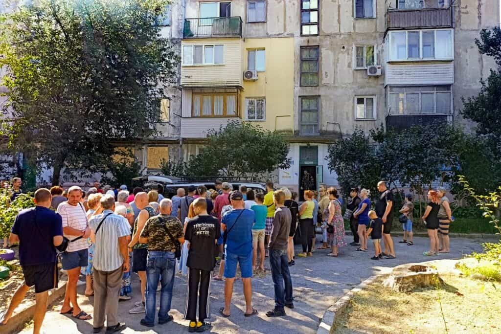 A group of people of various ages gathering and conversing outside old apartment buildings. The buildings show signs of wear and age, with a mixture of balconies and windows. A tree shades part of the gathering, and a car is parked nearby.