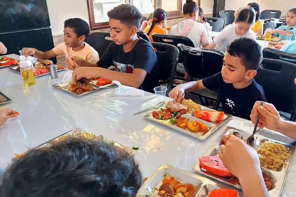 Several children seated at a long table, enjoying a meal with various dishes including rice, meat, and sliced watermelon. They are focused on their plates, using utensils to serve and eat.