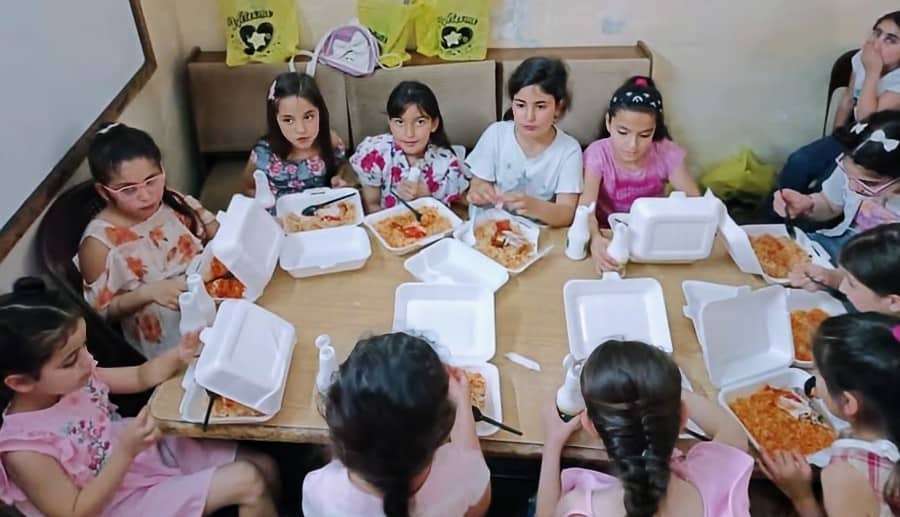 A group of young girls seated around a wooden table, each holding white styrofoam containers filled with food. They wear various outfits, from dresses to t-shirts, and some have their hair adorned with bows and hairbands.