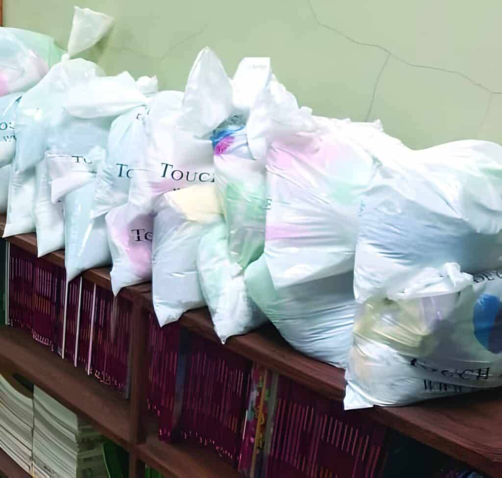 A line of large plastic bags filled with various food items, resting on top of a wooden bookshelf filled with rows of books. The bags have a printed text that includes 'Touch A Life'