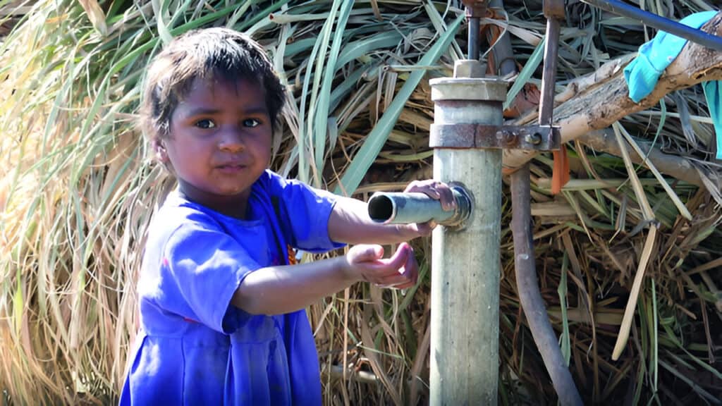 A young child in a blue shirt stands in front of a thatched background, holding onto a rustic metal water pump. The child looks directly at the camera with a hint of a smile, and their hand is on the pump handle, suggesting they may be about to draw water. The environment appears rural with natural lighting highlighting the scene.