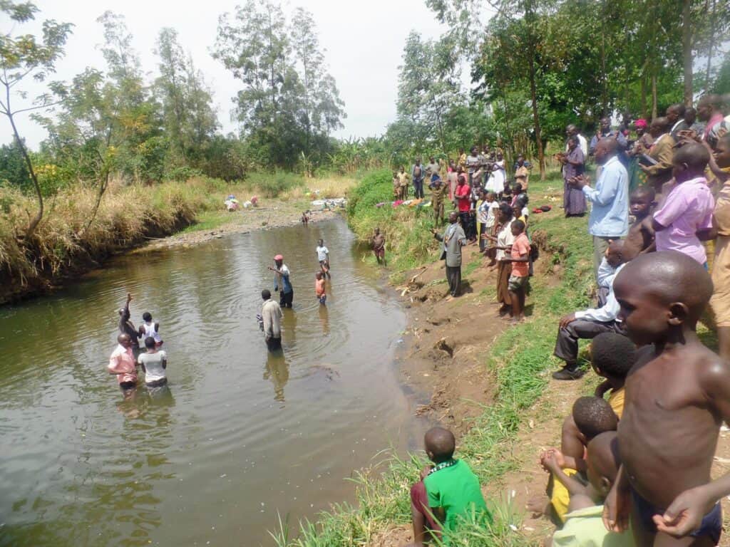 A congregation gathers by a Kenyan river, witnessing a baptism as individuals are immersed in the water, with onlookers standing along the riverbank.