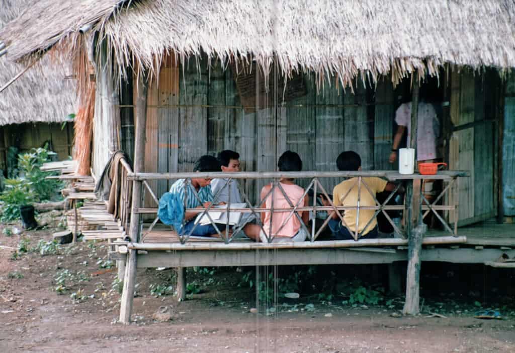 A group of young Akha men sit focused on their studies at a wooden table on the porch of a traditional thatched house, preparing for ministry work.