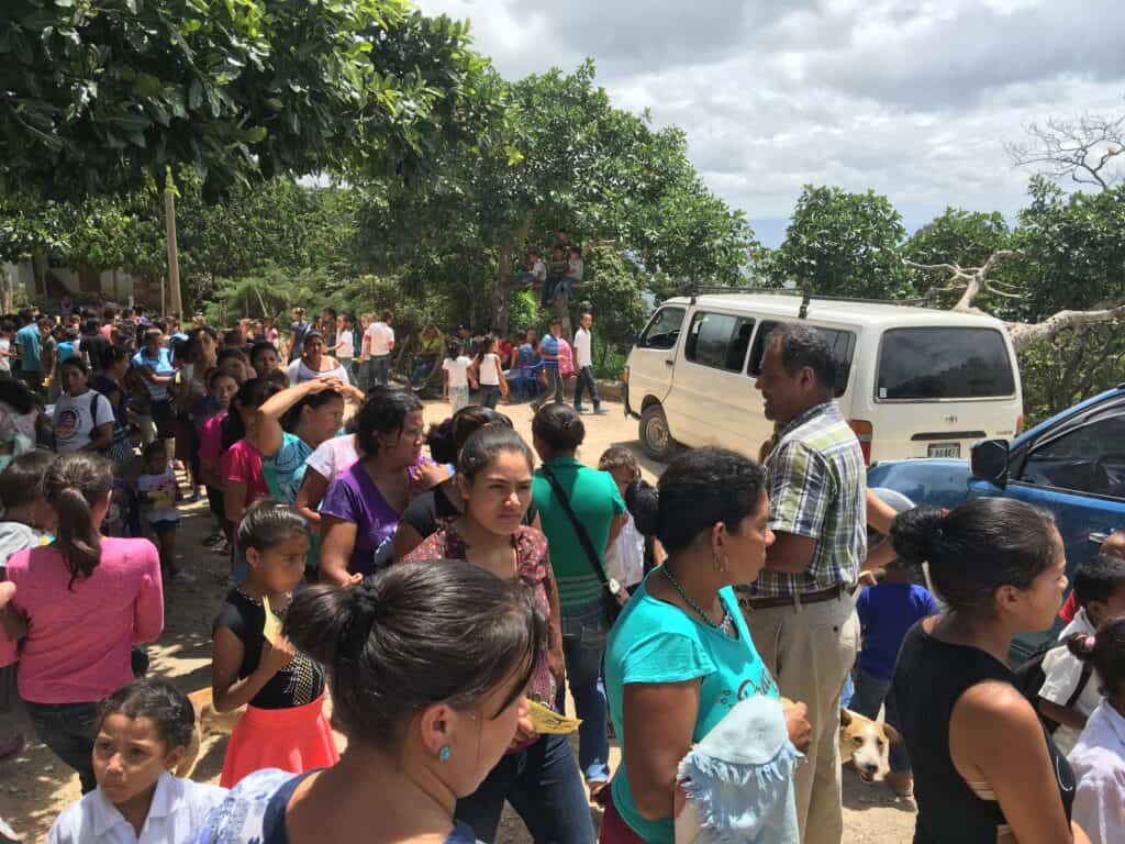 A bustling outdoor congregation in Zuzular, Honduras, with people of all ages gathering under the shade of trees, alongside parked vehicles.