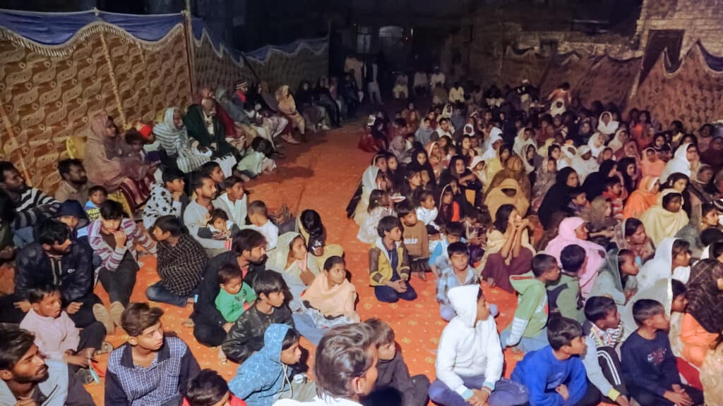 A large gathering of villagers, including men, women, and children, seated closely together on the ground, participating in an evangelism event