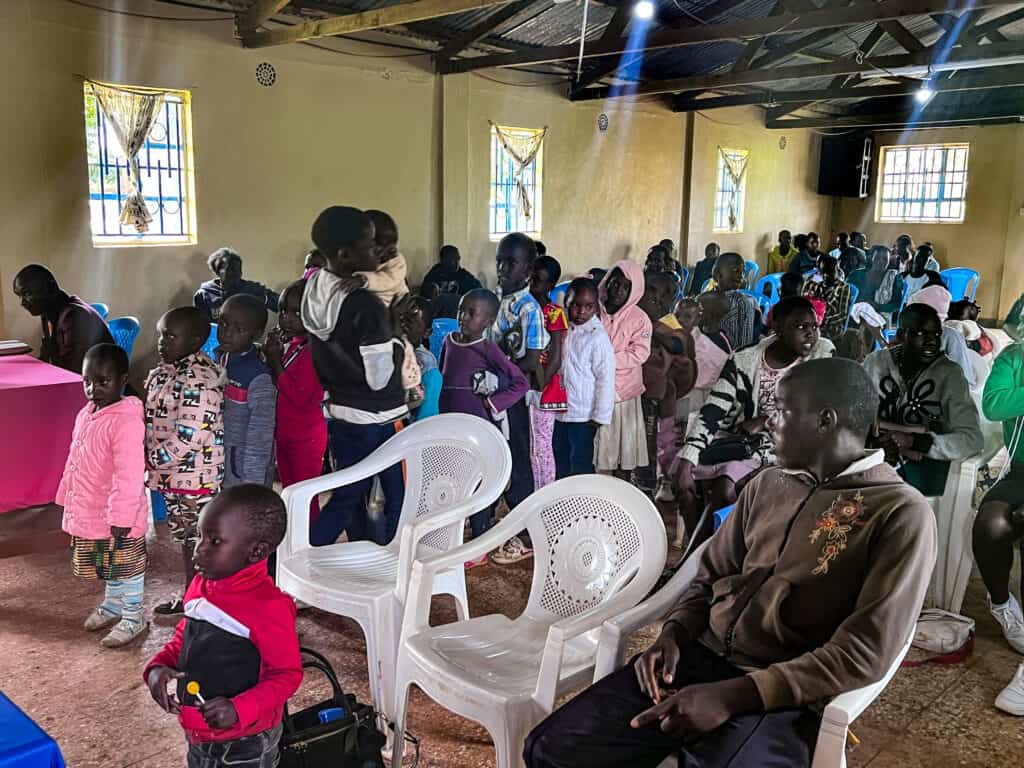 A room filled with children and adults, some standing and others seated, all attending an event at a community center.