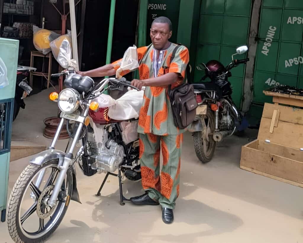 A pastor in traditional attire stands with a new motorbike, a testament to the support that aids his ministry's mobility and reach.