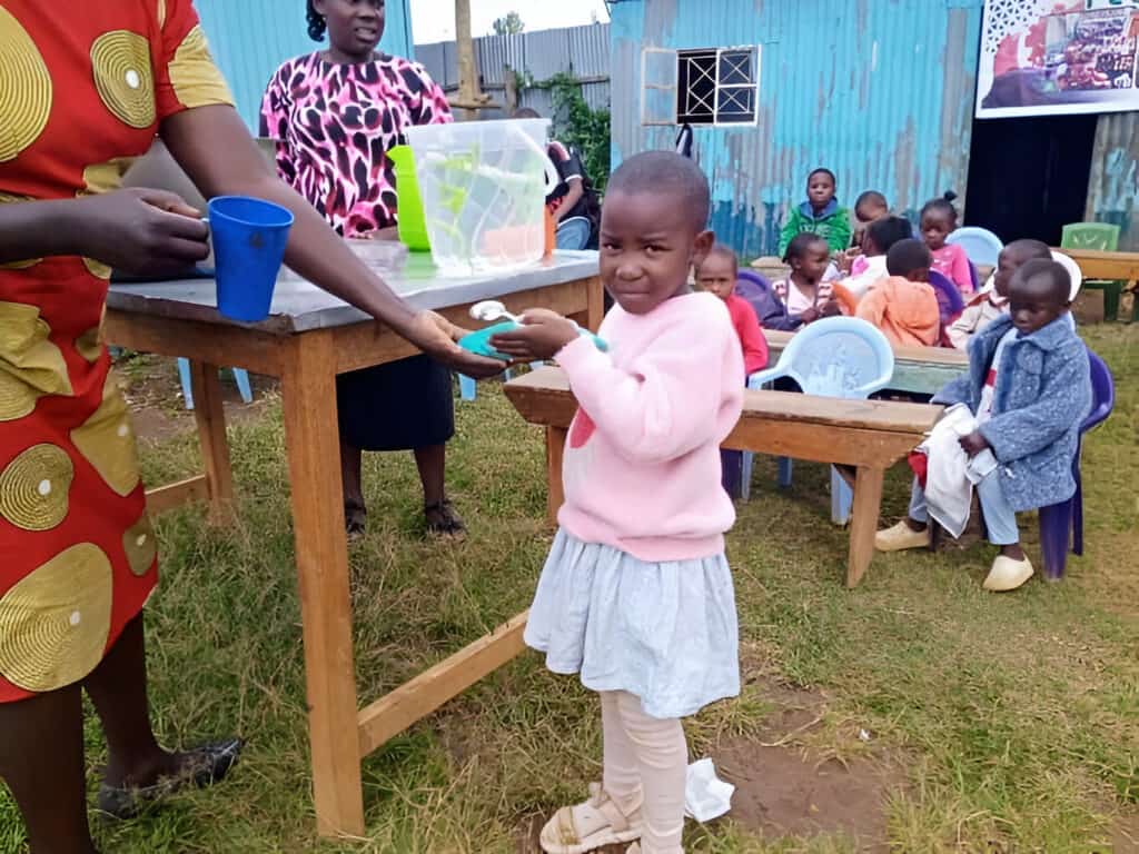 A young child receiving a cup of water at an outdoor community feeding center, with other children seated in the background.