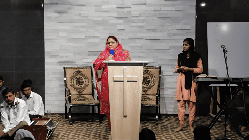 Mehwish stands at a podium, sharing her testimony in a church setting, with attendees listening intently.