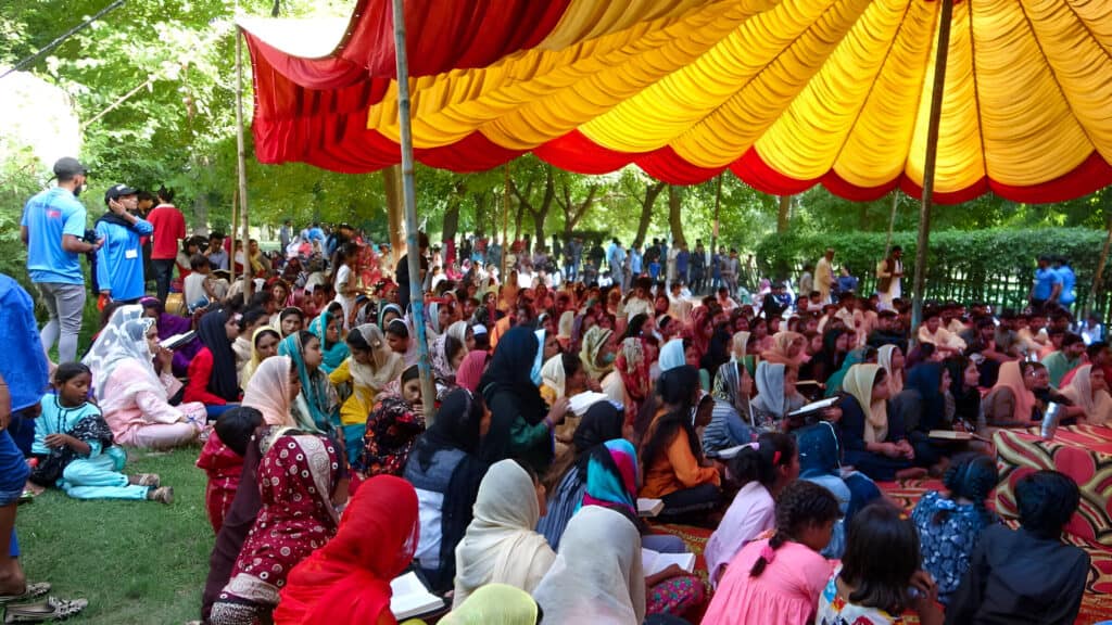 A diverse gathering of individuals, predominantly women and children, seated attentively under a vibrant red and yellow canopy in a park, engaged in a community event or educational activity.