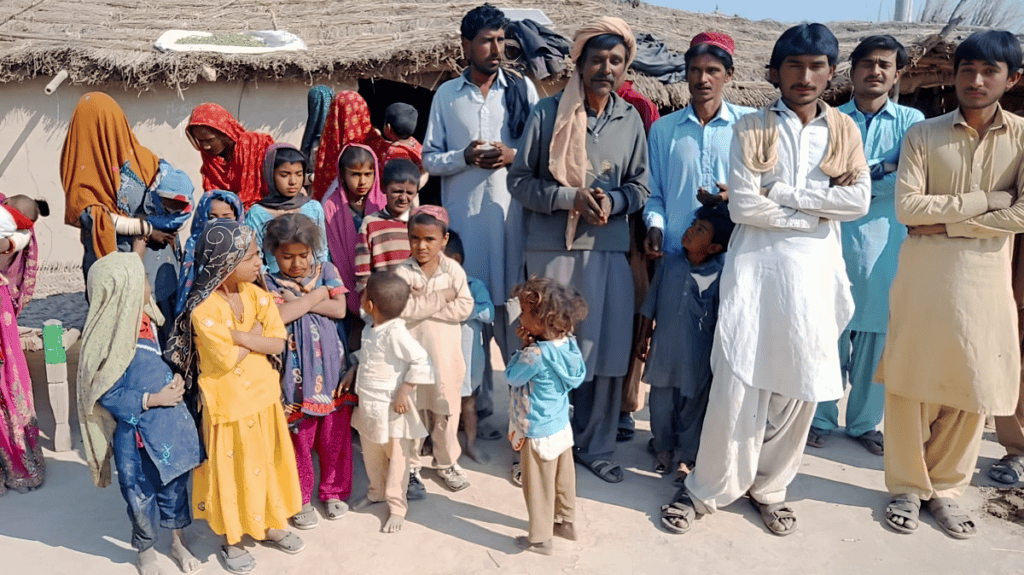 A gathering of Marwari individuals of all ages, displaying a mix of curiosity and resolve, stand before a traditional mud-block home, signifying a community on the cusp of spiritual transformation.