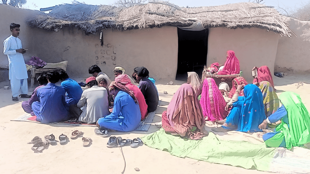 A young man preaches to an audience of villagers seated on mats in front of a traditional house, portraying a moment of communal learning and spiritual growth.