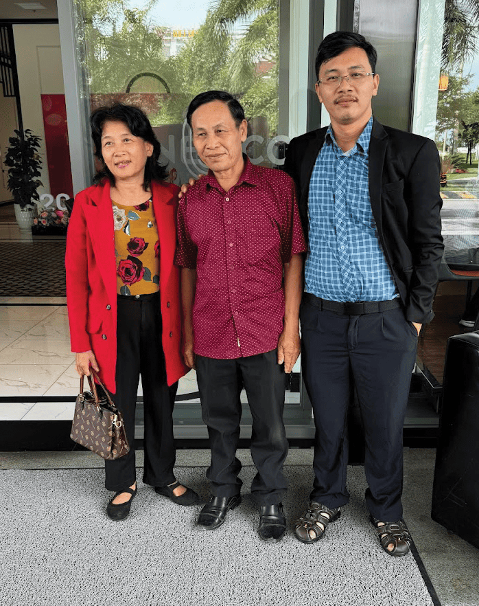 Three Vietnamese people standing together outside a building - a woman in a red coat, a middle-aged man in a purple shirt, and a younger man in a black suit with a blue checked shirt.