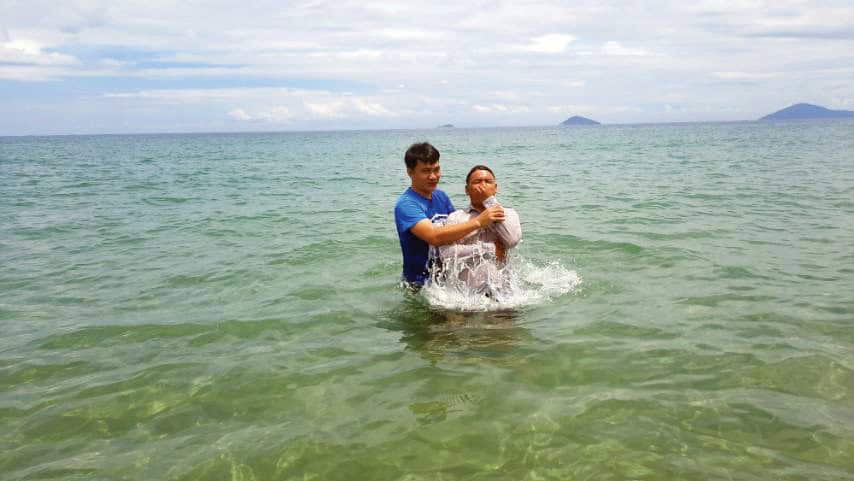 A person in a blue shirt baptizing another individual in clear turquoise ocean water. The baptism is taking place with the person being immersed while waves splash around them. Mountains or islands can be seen on the horizon under a partly cloudy sky.