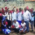 A group of new believer, each with their new Bible raised in the air having just received them, standing outside infront of a building.
