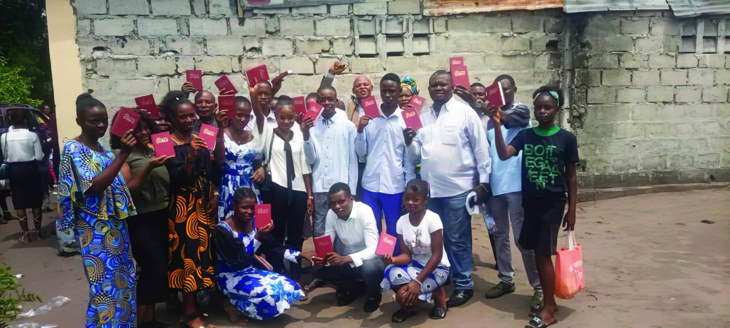 A group of new believer each with their new Bible raised in the air having just received them standing outside infront of a building