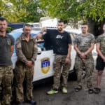 A group of six soldiers in the Ukraine in casual and camouflaged attire standing in front of a white car with a blue and yellow emblem. The environment is an outdoor setting with trees and a rustic wall in the background.