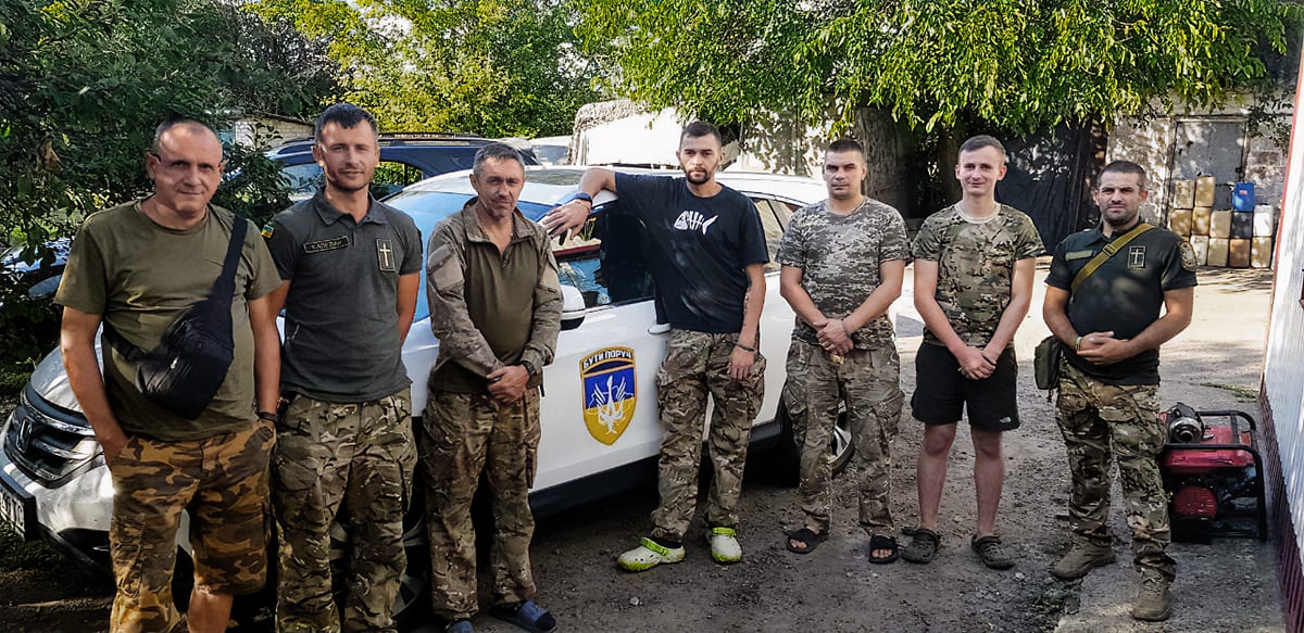 A group of six soldiers in the Ukraine in casual and camouflaged attire standing in front of a white car with a blue and yellow emblem. The environment is an outdoor setting with trees and a rustic wall in the background.