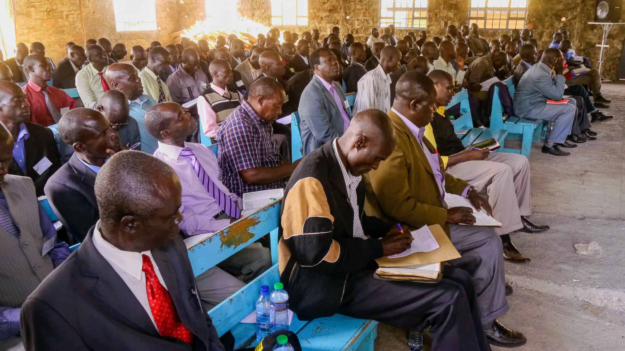 A group of Kenyan pastors sits attentively in a church, participating in a training session with open notebooks and Bibles, engaged in learning and discussion.