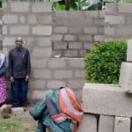 An elderly couple, with the woman wearing a vibrant patterned dress and the man in a dark shirt, stand in front of an unfinished brick structure surrounded by greenery. Nearby, there are stacks of concrete bricks.