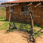 A makeshift soccer goal created ingeniously from discarded black irrigation tubes woven tightly across a wooden frame. Set against the backdrop of a brick house in a rural setting, the goal symbolizes the innovation and resourcefulness of local Honduran children, transforming agricultural waste into a playfield essential. The ground, compacted earth, bears evidence of frequent use, showcasing the passion and dreams of young football enthusiasts.