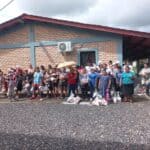 A group of adults and children standing infront of a brick building in Honduras. They are all looking at the camera, some are holding bags of food while others have the bags of food at their feet. Several of the adults are holding umbrellas to protect themselves from the sun.