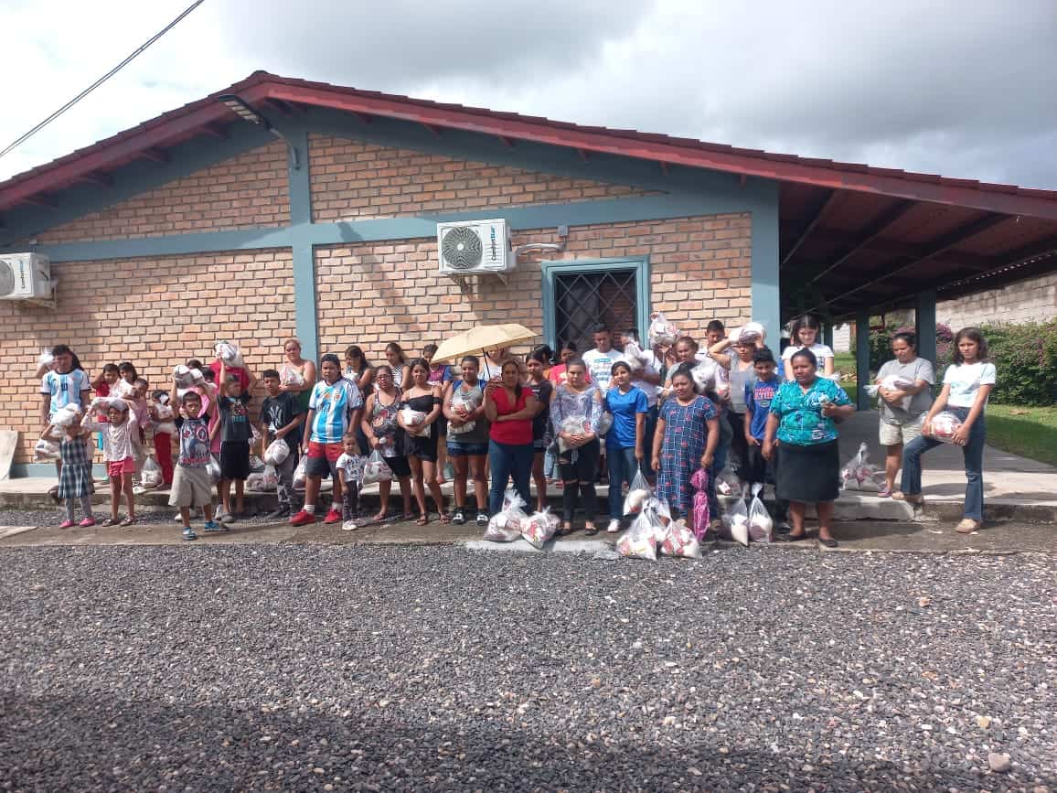 A group of adults and children standing infront of a brick building in Honduras. They are all looking at the camera, some are holding bags of food while others have the bags of food at their feet. Several of the adults are holding umbrellas to protect themselves from the sun.