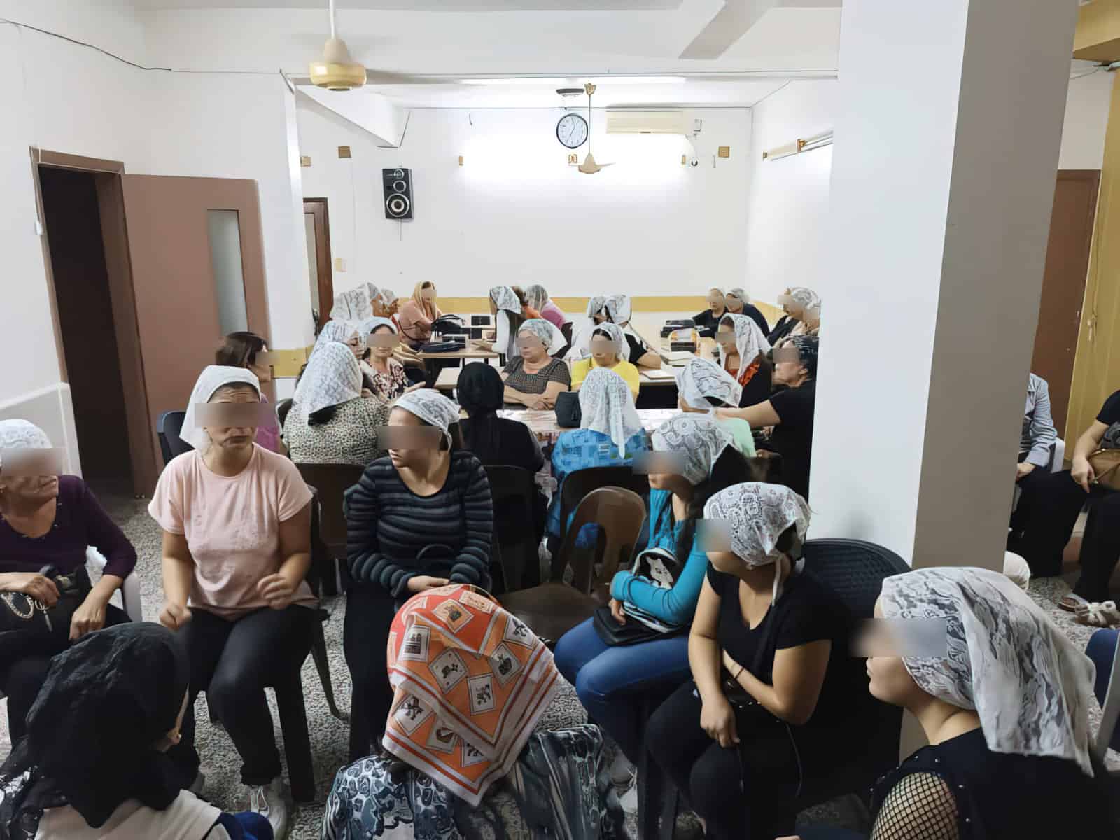 Murshid ladies (who were once unreached), approximately 30, who have already been evangelized sit in a white walled room in charis, some at tables in midst conversation with on another.