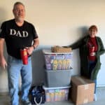 Dale and Cindee Dupaquier standing proudly in a garage next to a large storage bin and boxes filled with vitamin bottles for children in need.