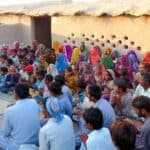 An outdoor gathering of a Pakistani church community in a desert setting, with members of all ages sitting on the ground, joyfully listening to the preached Word of God.
