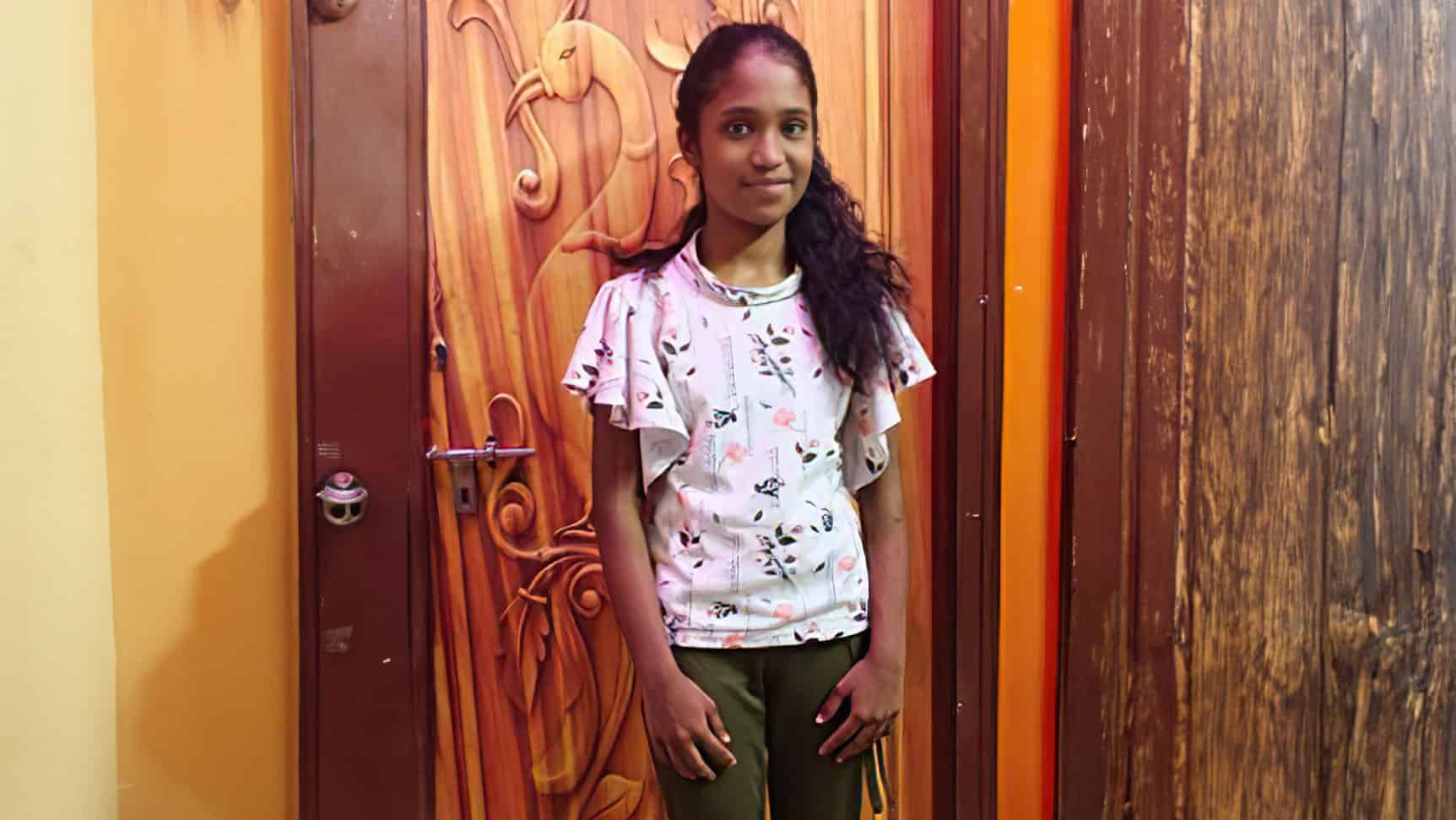 Sanjana, a young girl with a gentle smile stands confidently in front of a colorful wooden door adorned with intricate carvings.