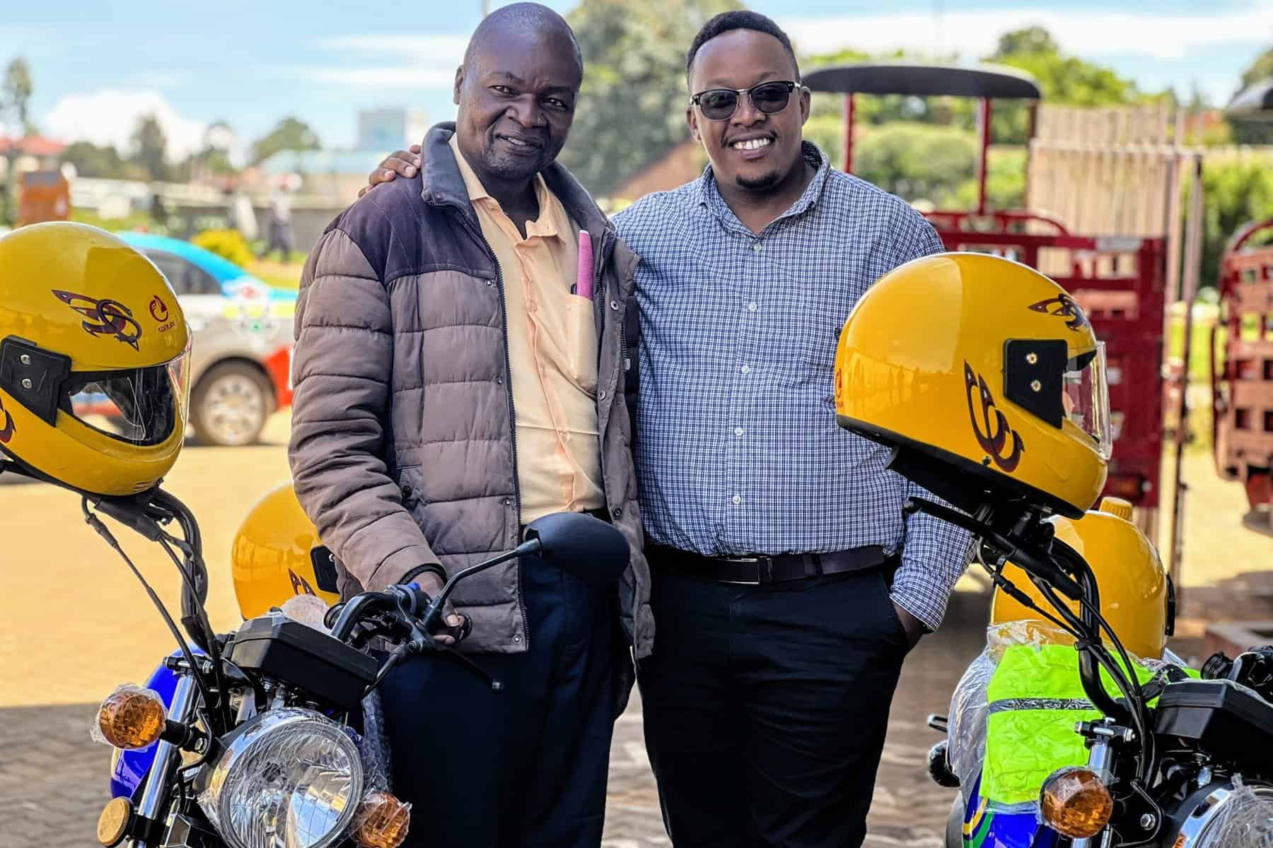Two smiling men standing next to new yellow motorbikes, representing the generous donation that aids pastors in their community work.