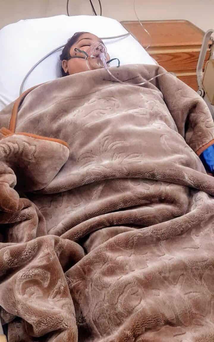 A patient is resting in a hospital bed covered with a blanket, receiving oxygen through a mask, suggesting a moment of medical care and recovery.