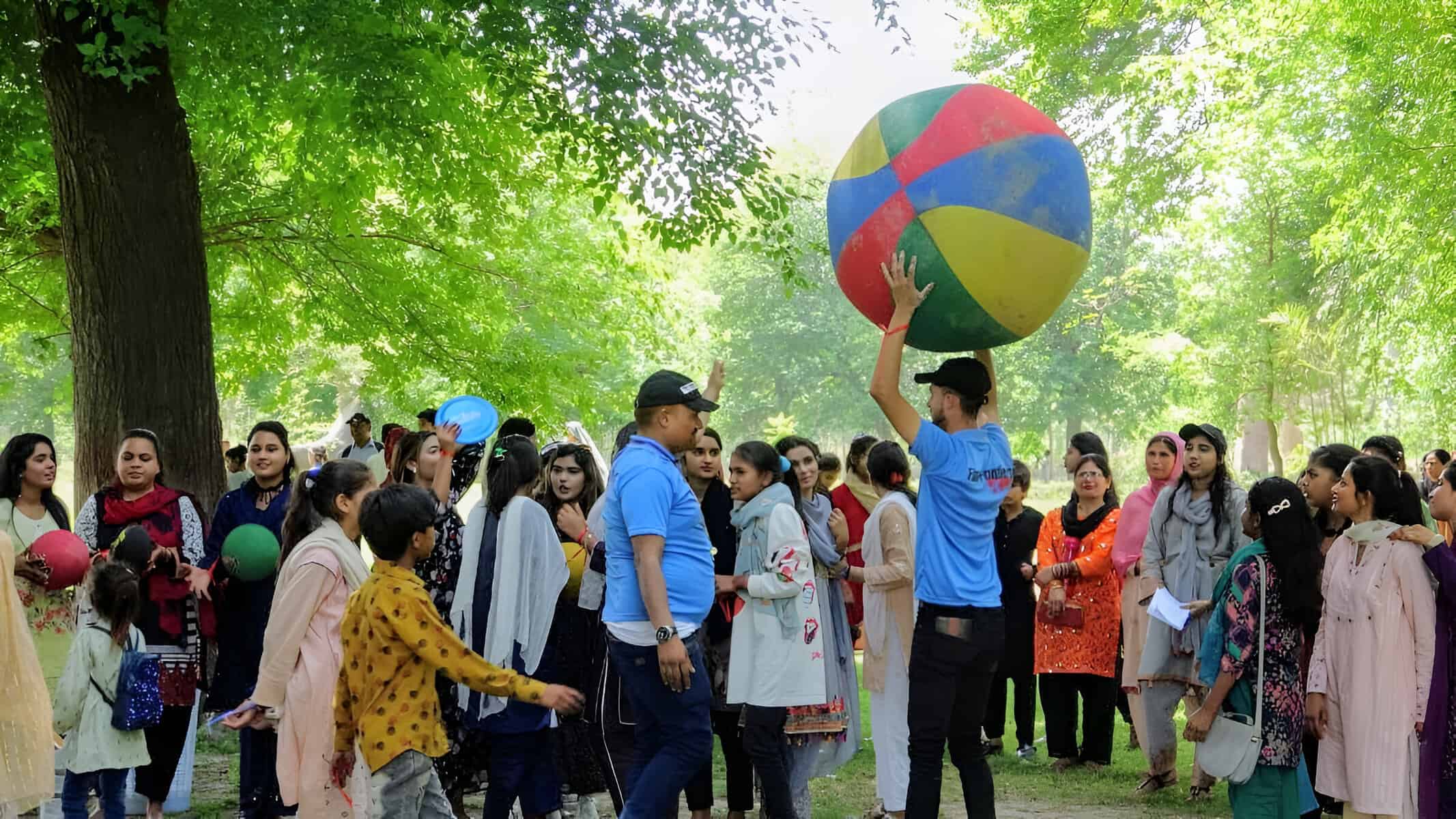 A group of joyful young people and adults engaging in outdoor activities, with a focus on a colorful oversized beach ball being tossed in the air among a crowd of smiling faces in a lush park setting.