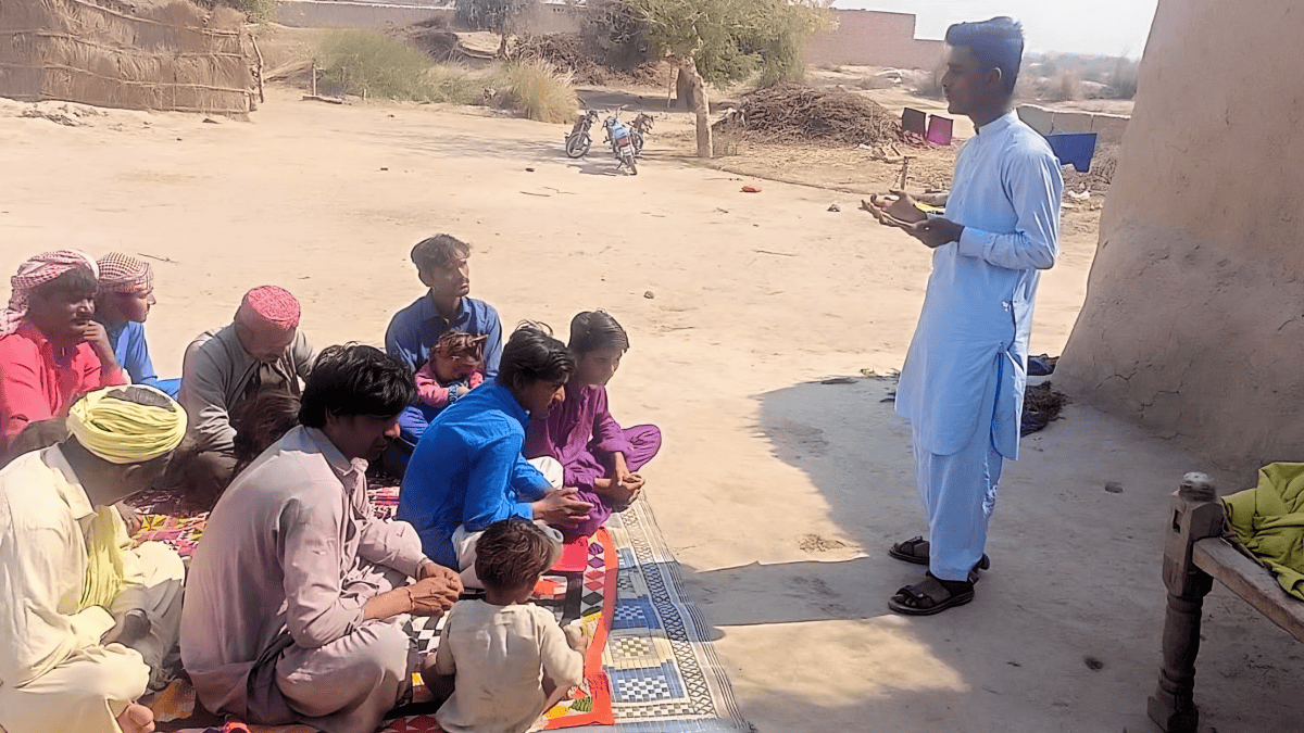 A young man stands teaching a captivated audience seated on a colorful mat in a sandy village setting, with motorcycles in the background symbolizing the journey they have undertaken.