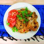 A white plate of spiced rice with chicken, garnished with tomato slices and fresh cilantro, set on a blue patterned cloth