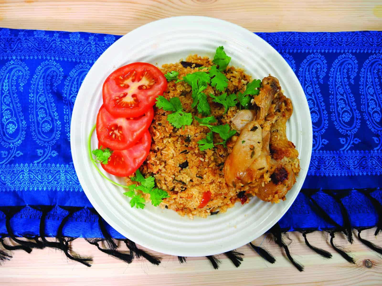 A white plate of spiced rice with chicken, garnished with tomato slices and fresh cilantro, set on a blue patterned cloth