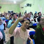 A joyful gathering of Vietnamese church members with raised hands and smiling faces in a simple green-walled room with ceiling fans.