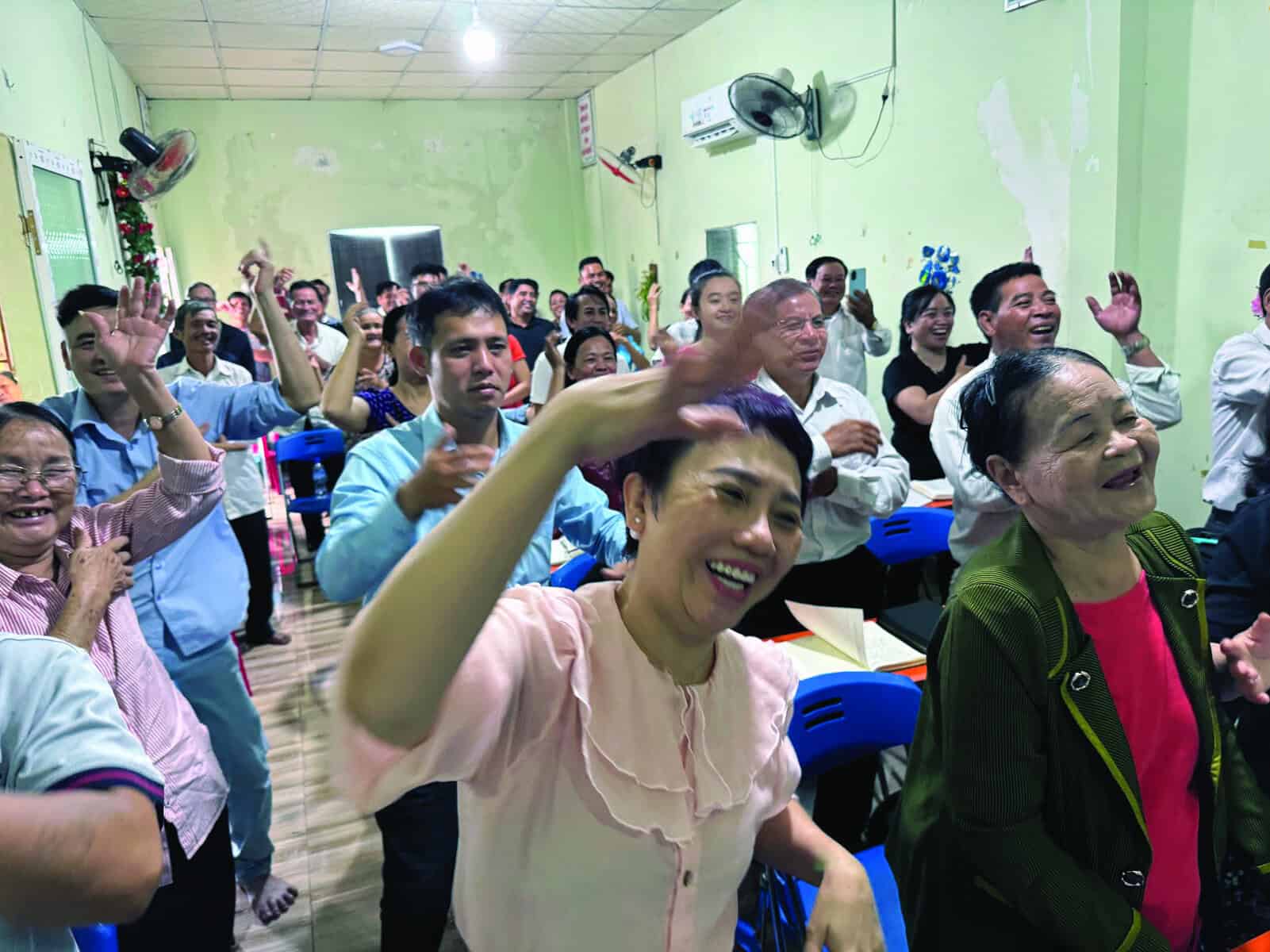 A joyful gathering of Vietnamese church members with raised hands and smiling faces in a simple green-walled room with ceiling fans.