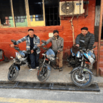 Three men stand next to their new motorcycles outside of a brick building in Nepal