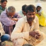 A man has his eyes closed while squating on the sand. Around him are several other people, women and men, who are also sitting with their eyes closed as they pray. In the background you see the side of a sand hut building and a motorcycle.