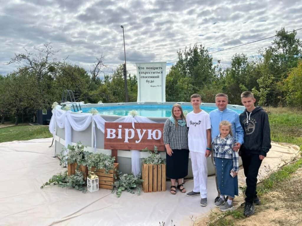 A family stands beside a baptism pool decorated with flowers and a sign reading віpую (I believe)